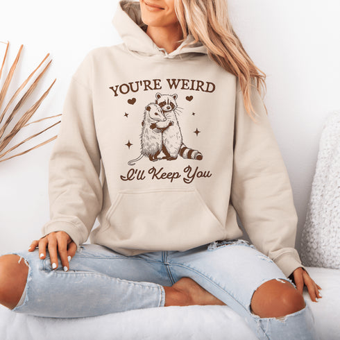 Celebrate the beauty of quirky connections with our comfy hoodie, featuring an adorable illustration of a raccoon and possum sharing a heartwarming hug.